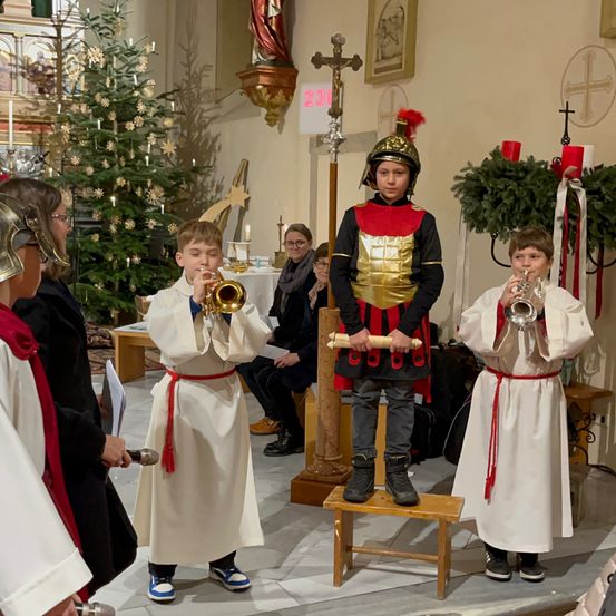 Mehrere Kinder in weißen Gewändern und roten Schärpen spielen Trompeten, eines steht auf einem Holzhocker in einer goldenen und roten Kostümierung und einem Helm. Dahinter sind ein Weihnachtsbaum und ein Kreuz zu sehen.