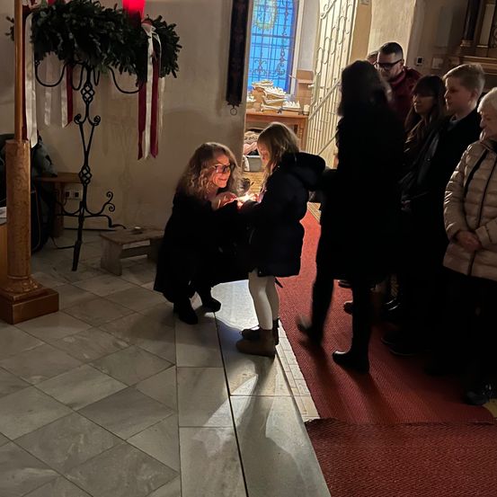 Eine Frau hilft einem jungen Mädchen, eine Kerze in einer Kirche anzuzünden, während Leute zusehen.