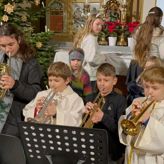 Mehrere Kinder in weißen Gewändern spielen Trompeten bei einer kirchlichen Veranstaltung. Ein geschmückter Weihnachtsbaum und eine Statue sind im Hintergrund zu sehen.