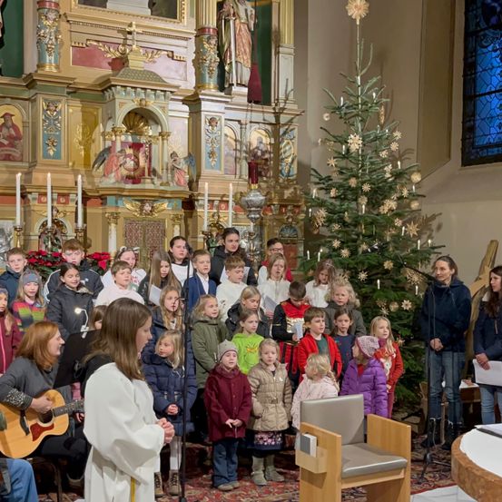 Ein Kinderchor steht vor einem geschmückten Weihnachtsbaum in einer Kirche, begleitet von einer Frau, die Gitarre spielt. Dahinter stehen Erwachsene und singen, mit einem Holzstuhl und einem Tisch in der Nähe.