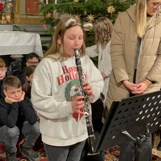 Ein Mädchen in einem Feiertags-Sweatshirt spielt Klarinette bei einer Weihnachtsveranstaltung, während mehrere Kinder zusehen und Erwachsene in der Nähe sind.