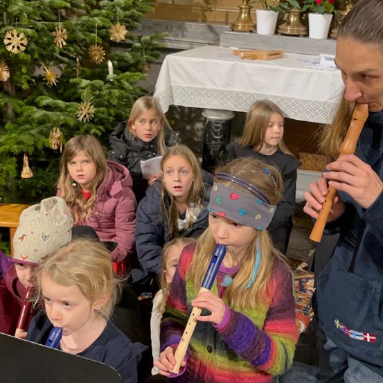 Eine Gruppe von Kindern spielt Holzflöten in einer Kirche, angeführt von einer Frau mit einer Flöte, mit einem Weihnachtsbaum und Kerzen im Hintergrund.