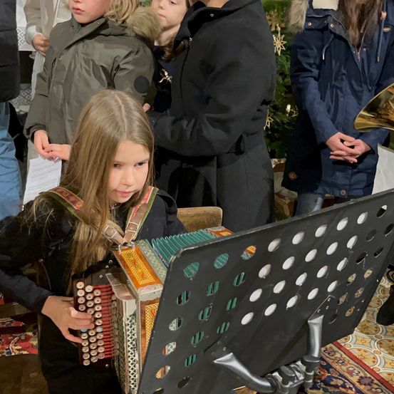 Ein junges Mädchen spielt Akkordeon bei einer Zusammenkunft, trägt eine schwarze Jacke. Hinter ihr sind Menschen versammelt, einer hält ein Blatt Papier. Ein Weihnachtsbaum und ein Posaune sind ebenfalls anwesend.