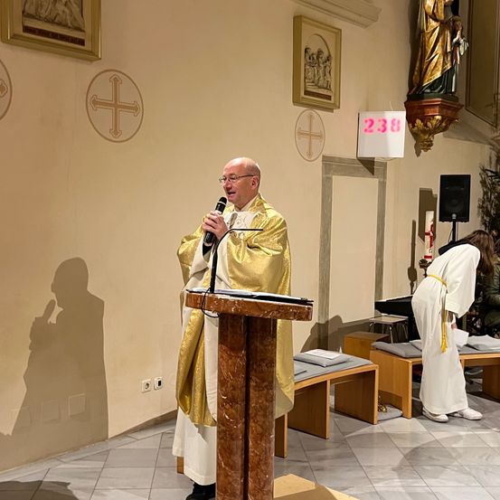 Ein Priester steht in einer Kirche an einem Podium mit Mikrofon, trägt eine goldene und weiße Robe, während eine andere Person in Weiß in der Nähe kniet.