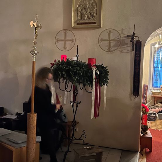 Eine Kircheninnenansicht mit einem geschmückten Altar, einem Kreuz auf einem Ständer und einer Frau, die Gegenstände ordnet. An der Wand befindet sich ein gerahmtes Bild, Kreuze und ein Fenster mit blauen Lichtern.