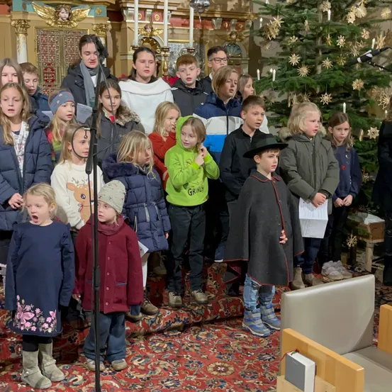 Ein Kinderchor steht in einer Kirche und singt vor einem geschmückten Weihnachtsbaum und einem erwachsenen Menschen in der Nähe.