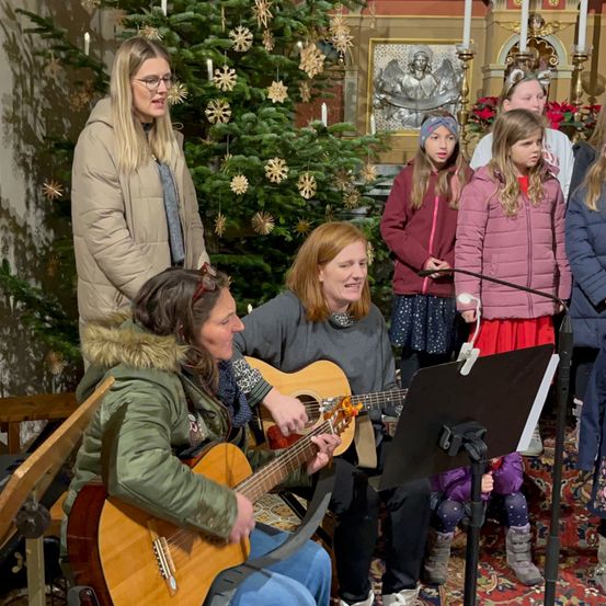 Eine Gruppe von Menschen singt Weihnachtslieder in einer Kirche. Eine Frau spielt Gitarre, während andere hinter ihr stehen, darunter Kinder in Winterkleidung. Ein Weihnachtsbaum ist mit Ornamenten geschmückt.