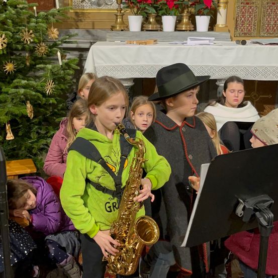 Ein junges Mädchen spielt Saxophon, während andere zusehen. Ein Weihnachtsbaum und ein Kirchenhintergrund sind sichtbar.