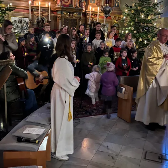 Eine Gruppe von Menschen, einschließlich Kinder, versammelt sich in einer Kirche, einige halten Kerzen und ein Priester steht vor einem Weihnachtsbaum. Ein Mikrofon steht auf einer Bank.