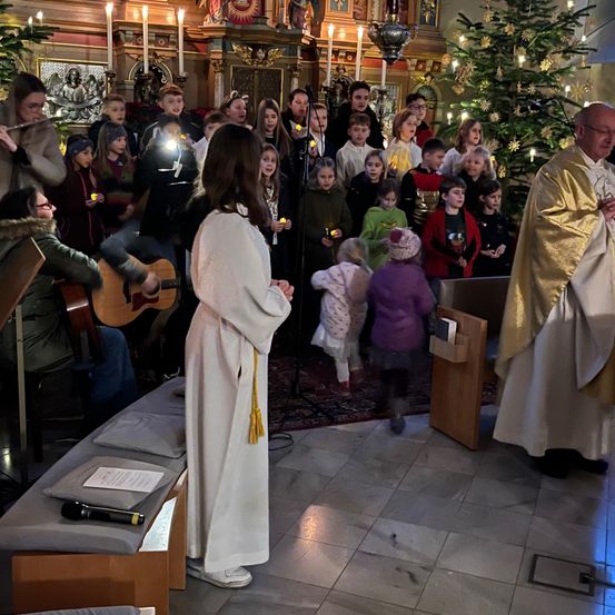 Eine Gruppe von Menschen, einschließlich Kinder, versammelt sich in einer Kirche, einige halten Kerzen und ein Priester steht vor einem Weihnachtsbaum. Ein Mikrofon steht auf einer Bank.