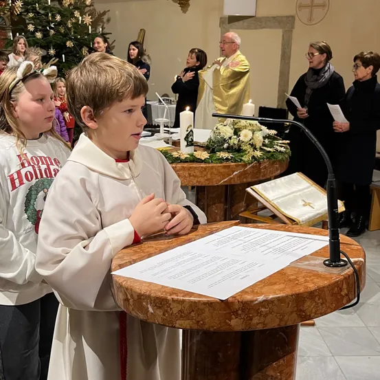 Ein Junge in einer weißen Robe liest ein Papier an einem Podest, während andere hinter ihm stehen. Der Ort ist eine Kirche mit einem Weihnachtsbaum, Kerzen und einem Priester.