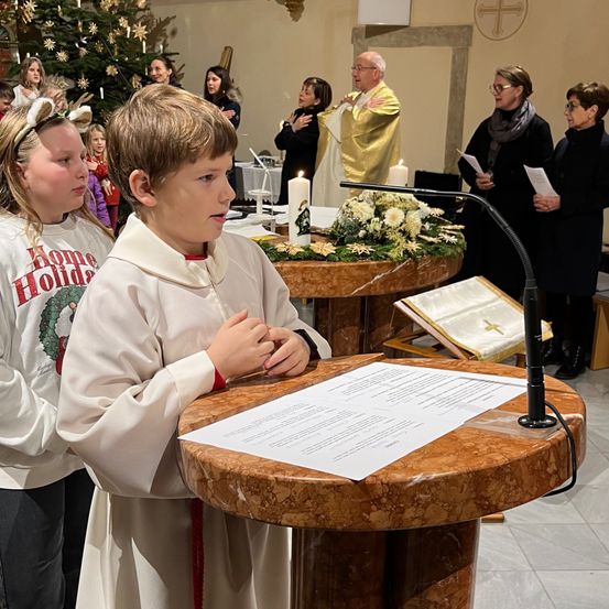 Ein Junge in einer weißen Robe liest ein Papier an einem Podest, während andere hinter ihm stehen. Der Ort ist eine Kirche mit einem Weihnachtsbaum, Kerzen und einem Priester.