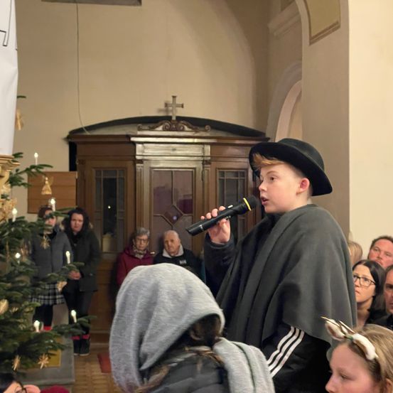 Ein Junge in einem schwarzen Hut und Umhang hält ein Mikrofon und singt in einer Kirche. Hinter ihm steht ein mit Kerzen geschmückter Weihnachtsbaum. Mehrere Menschen schauen zu, darunter ein älterer Mann mit Brille.