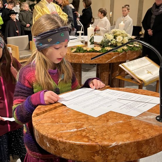 Ein kleines Mädchen mit Kopfband liest ein Papier am Altar einer Kirche, umgeben von Menschen und Kerzen.