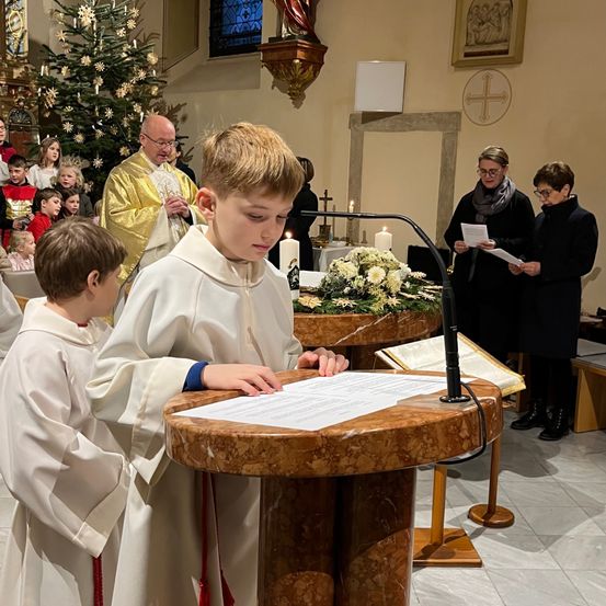 In einer Kirche liest ein junger Junge von einem Skript an einem Pult, während ein Priester und Kinder im Hintergrund stehen. Ein Weihnachtsbaum und Kerzen sind vorhanden.