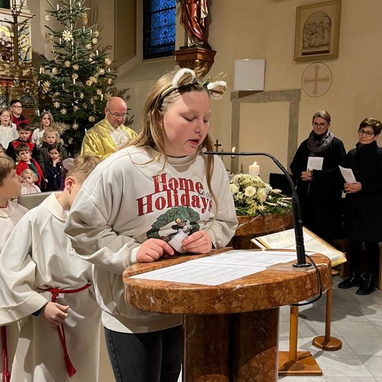 Ein junges Mädchen steht an einem Rednerpult und hält eine Rede in einer Kirche während eines Feiertagsgottesdienstes. Hinter ihr stehen mehrere Kinder in weißen Gewändern. Ein Weihnachtsbaum und Kerzen befinden sich auf der linken Seite.