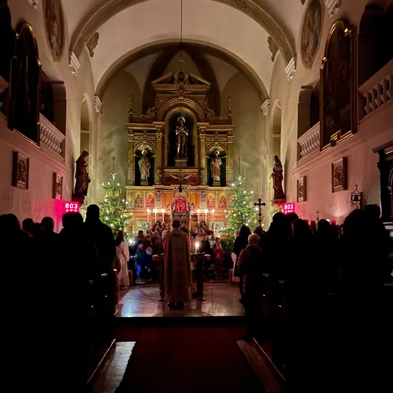 Ein Priester leitet einen Gottesdienst in einer Kirche, wobei Menschen auf den Kirchenbänken sitzen, Kerzen auf dem Altar brennen und mit Lichtern geschmückte Weihnachtsbäume im Hintergrund stehen.