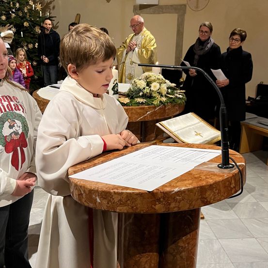 Ein Junge in einer weißen Robe steht an einem Pult und liest ein Papier. Dahinter steht ein Priester in goldener Robe, der aus einem Buch liest. Im Hintergrund befinden sich Zuschauer, darunter eine Frau und Kinder.
