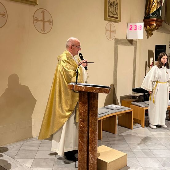 Ein Priester steht an einem Pult, hält ein Mikrofon in der Hand und hält eine Predigt in einer Kirche. Ein Mädchen in einem weißen Gewand steht in der Nähe.