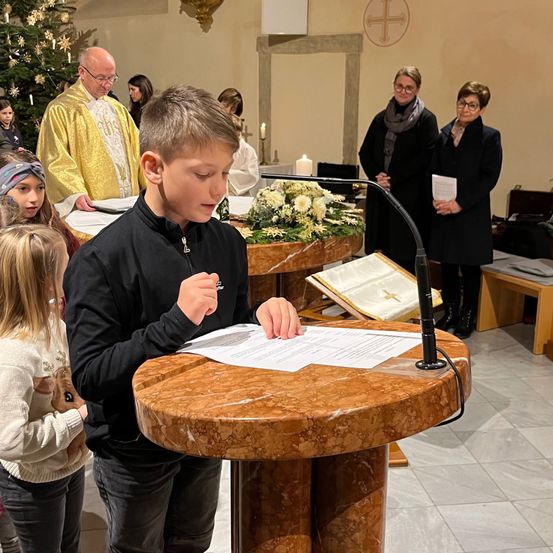 Ein junger Junge liest laut aus einem Blatt Papier an einem Podium während eines Gottesdienstes. Hinter ihm steht ein Priester am Altar, und zwei Frauen schauen zu.
