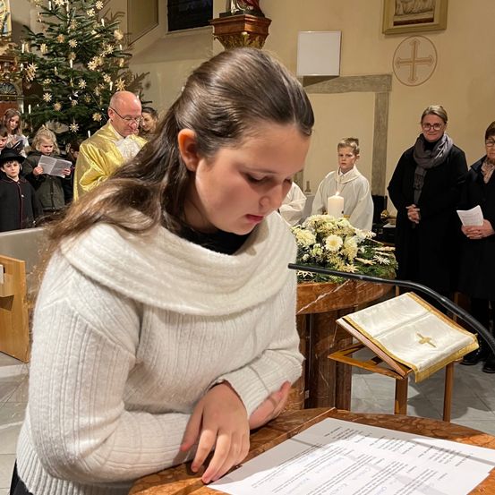 Ein Mädchen mit braunen Haaren kniet vor einem Kirchenaltar und schaut auf ein Stück Papier. Hinter ihr stehen Menschen vor einem Weihnachtsbaum und einer Kerze.
