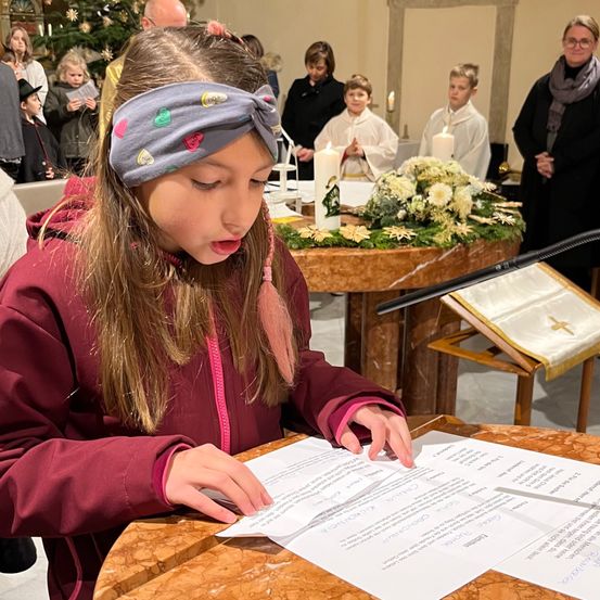 Ein Mädchen liest während eines Gottesdienstes ein Dokument, mit Kerzen und einer Blumendekoration hinter ihr. Andere Menschen, einschließlich Kinder, stehen und beobachten.
