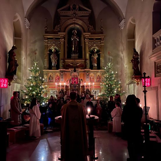 Eine Gruppe von Menschen versammelt sich in einer Kirche mit zwei Weihnachtsbäumen und einem Priester, der einen kerzenbeleuchteten Gottesdienst leitet.