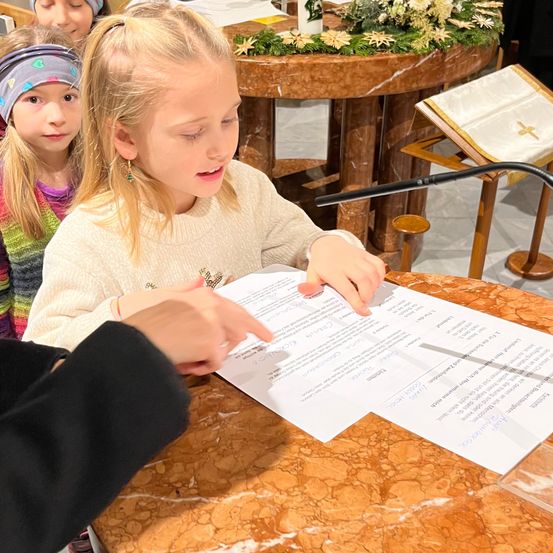 Ein junges Mädchen liest ein Dokument an einem Tisch in einer Kirche mit einem Mikrofon. Ein weiteres Mädchen steht hinter ihr und schaut zu.