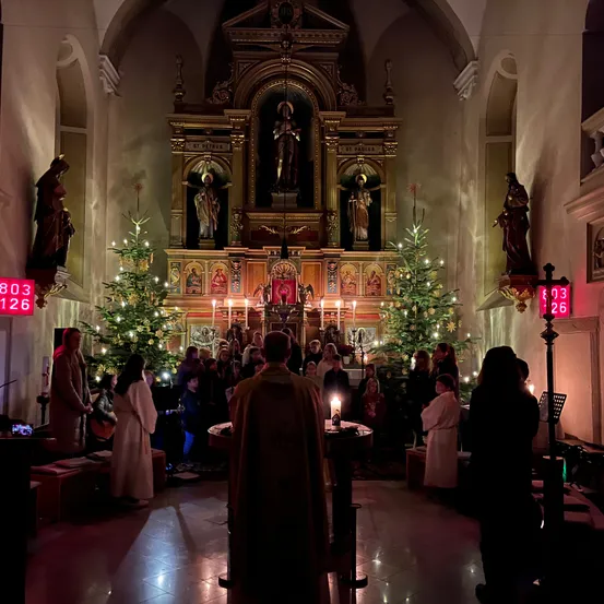 Eine Menschenmenge versammelt sich in einer Kirche mit Weihnachtsbäumen und Kerzen. Ein Priester steht vor einem Altar mit brennenden Kerzen. Die Zahlen 803 und 126 sind auf Schildern angebracht.