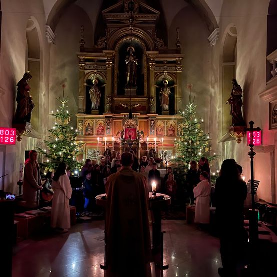 Eine Menschenmenge versammelt sich in einer Kirche mit Weihnachtsbäumen und Kerzen. Ein Priester steht vor einem Altar mit brennenden Kerzen. Die Zahlen 803 und 126 sind auf Schildern angebracht.