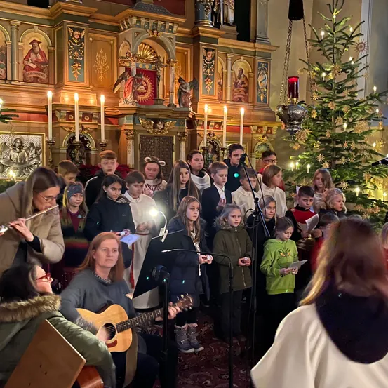 Ein Kinderchor in einer Kirche singt Weihnachtslieder mit einer Chorleiterin, die die Flöte spielt, umgeben von Erwachsenen und einem geschmückten Weihnachtsbaum.
