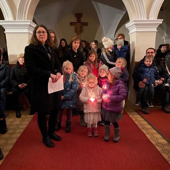 Eine Gruppe von Kindern, die brennende Kerzen in einer Kirche halten, angeführt von einer Frau mit einem Papier. Im Hintergrund sitzen Menschen auf Bänken.