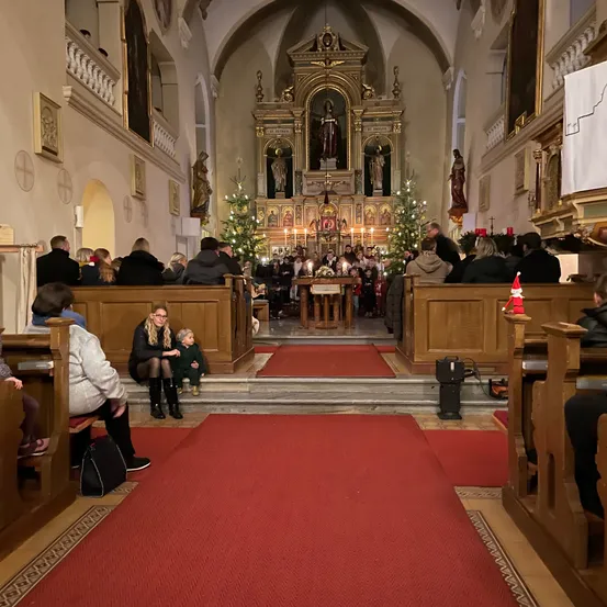 Eine Innenansicht einer Kirche mit Menschen auf Kirchenbänken. Der Altar ist mit Weihnachtsbäumen und Statuen geschmückt. Rote Teppiche führen zum Altar.