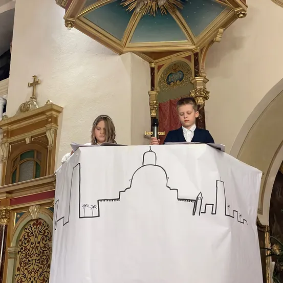 Zwei Kinder stehen in einer Kirche an einem Podest mit einer Zeichnung einer Stadt auf einem weißen Hintergrund. Der Junge hält ein Mikrofon, während das Mädchen auf ein Stück Papier schaut. Der Podest ist mit goldenen Akzenten und einer kunstvoll gestalteten Decke verziert.