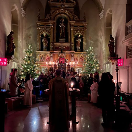 Eine Gruppe von Menschen in einer Kirche, die um einen Priester an einem Altar herumstehen. Kerzen und Weihnachtsbäume schmücken den Raum.