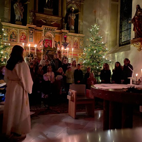 Eine Gruppe von Menschen, einschließlich Kindern, versammelt sich in einer Kirche und singt mit einem Weihnachtsbaum und Kerzen. Eine Frau in einem weißen Gewand steht vor ihnen.