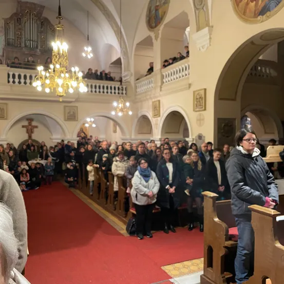 Eine große Gruppe von Menschen steht in Kirchenbänken in einer Kirche. Ein roter Teppich führt zum Vordersitz, wo eine Person steht. Andere sitzen auf dem Boden. Kronleuchter hängen von der Decke.