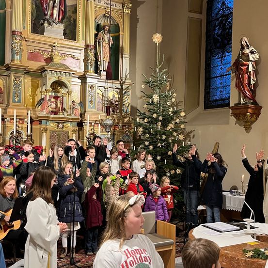 Ein Kinderchor singt in einer Kirche, mit einem Altar, Weihnachtsbaum und Statuen im Hintergrund.