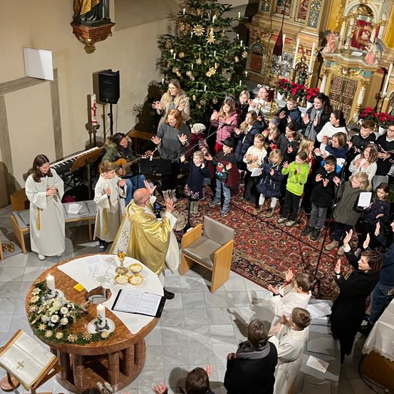 Eine Gemeinde versammelt sich in einer Kirche für einen Gottesdienst, mit einem Priester am Altar, Kindern davor und einem Weihnachtsbaum im Hintergrund.