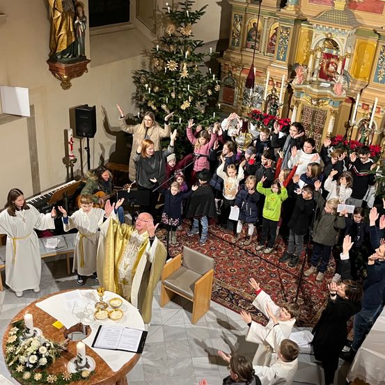 Ein Priester steht vor einem Kirchenaltar mit einem Chor von Kindern und Erwachsenen in weißen Gewändern. Die Kinder halten Kerzen und heben die Hände. Ein Weihnachtsbaum ist mit Lichtern und Blumen geschmückt.