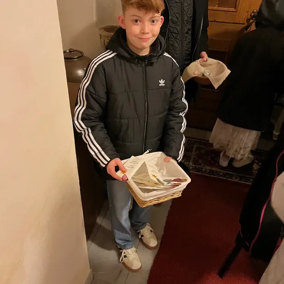 Ein Junge in einer schwarzen Adidas-Jacke mit weißen Streifen hält einen Korb mit Karten und Blumen in der Hand und steht in einem Korridor. Hinter ihm ist eine Person teilweise zu sehen, sowie ein hölzerner Schrank und ein Teppich.