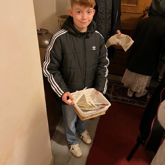 Ein Junge in einer schwarzen Adidas-Jacke mit weißen Streifen hält einen Korb mit Karten und Blumen in der Hand und steht in einem Korridor. Hinter ihm ist eine Person teilweise zu sehen, sowie ein hölzerner Schrank und ein Teppich.