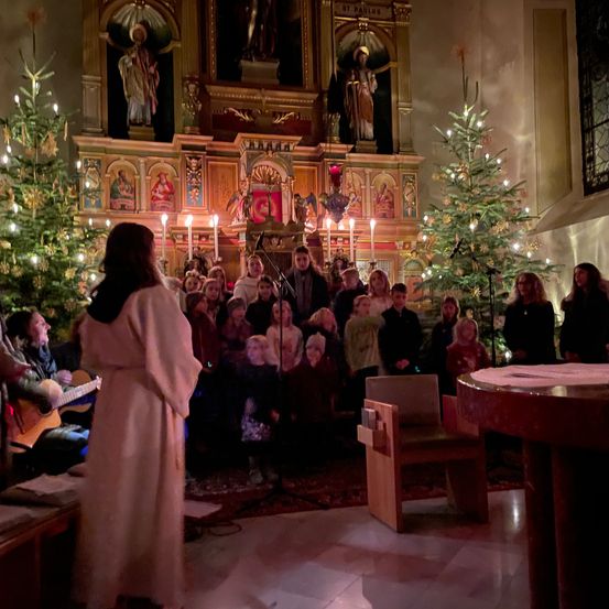 Ein Kinderchor singt in einer für Weihnachten geschmückten Kirche. Eine Frau spielt Gitarre in der Ecke.