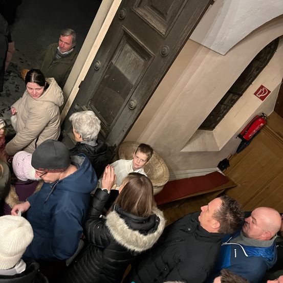 Eine Gruppe von Menschen in Winterkleidung versammelt sich um eine Tür in einer Kirche. Ein Feuerlöscher ist an der Wand neben der Tür montiert.
