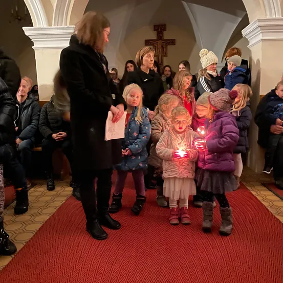 Mehrere Kinder und Erwachsene stehen mit Kerzen in einer Kirche. Eine Frau vorne hält ein Papier. Leute sitzen auf Bänken. Ein Kreuz hängt an der Wand.