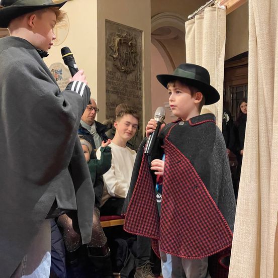 Zwei junge Jungen in Hüten und Umhängen halten Mikrofone, einer trägt einen schwarzen Hut und Umhang, der andere rot und schwarz. Ein Publikum sitzt hinter ihnen.