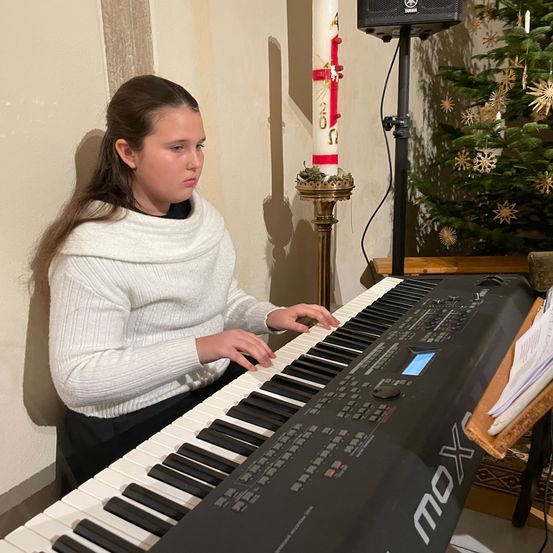 Ein junges Mädchen spielt Klavier in einer Kirche, mit einem Weihnachtsbaum und einer Kerze hinter ihr.