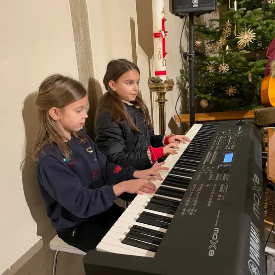 Zwei junge Mädchen sitzen vor einem Klavier, eine spielt, während die andere zuschaut. Hinter ihnen stehen ein Weihnachtsbaum und eine Kerze.