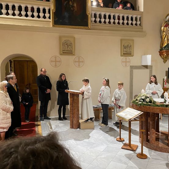 Eine religiöse Zeremonie in einer Kirche. Altardiener stehen neben einem geschmückten Altar. Zwei Erwachsene stehen bei einem Podest. Eine Statue eines Heiligen hängt oben.