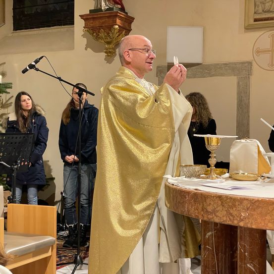 Ein Priester hält in einer Kirche eine Predigt. Er hält ein weißes Objekt in der Hand. Ein Mikrofon steht auf einem Ständer. Dahinter stehen Menschen, einer sitzt.